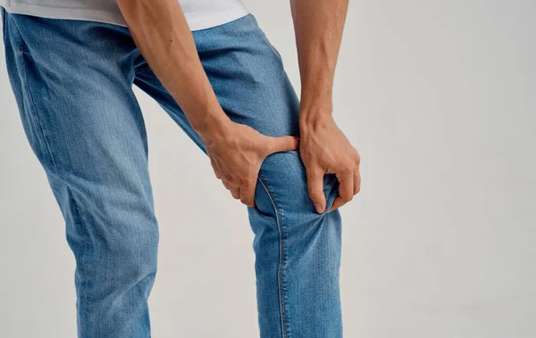 man in jeans touches his knee with his hands on a light background cropped view