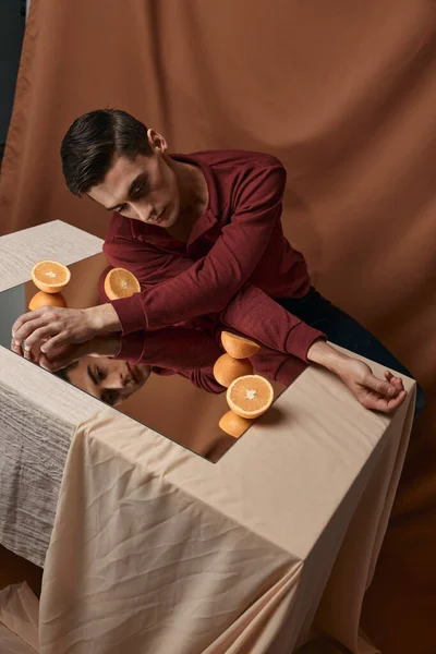 A guy in a shirt sits at a table with a mirror and orange oranges fabric background — Stock Photo, Image