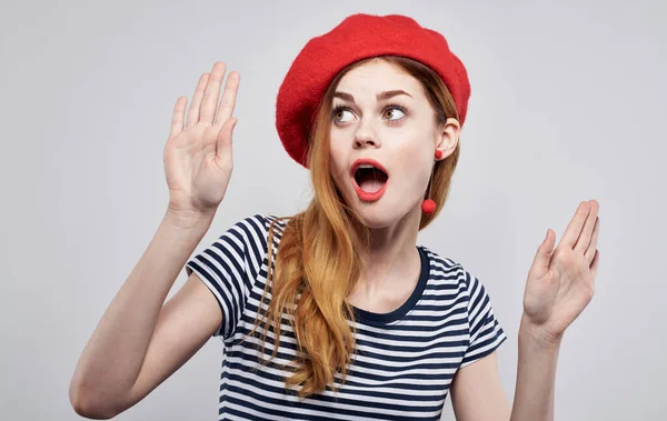 Fröhliche Frau mit rotem Hut und gestreiftem T-Shirt gestikuliert mit den Händen und lächelt verschränkt. — Stockfoto