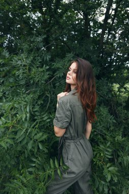 Ormandaki yeşil çalıların yanındaki tulumlu kadın yan yola bakıyor. 