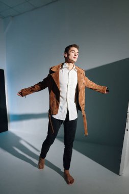 Kahverengi ceketli yakışıklı adam kollarını arka planda gri bir manzaraya doğru açıyor.