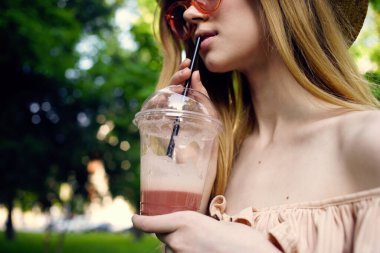 Bir bardak temiz hava içeceğiyle şapkalı ve gözlüklü neşeli kadın.