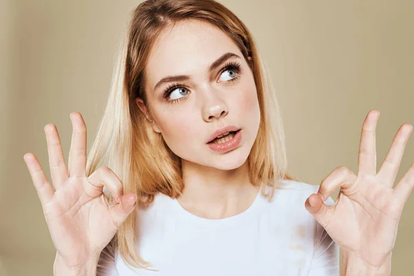 Femme blonde joyeuse dans un t-shirt blanc gestes avec ses émotions de la main fond beige — Photo