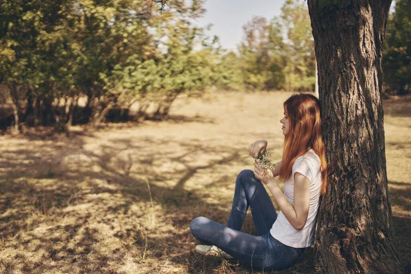 The traveler sits near a tree outdoors in the forest in jeans and a t-shirt — Stock Photo, Image