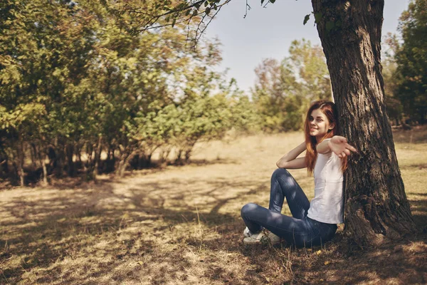 Ο ταξιδιώτης κάθεται κοντά σε ένα δέντρο έξω στο δάσος με τζιν και t-shirt — Φωτογραφία Αρχείου