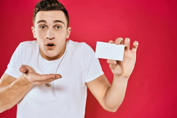 Een man in een wit T-shirt met een visitekaartje in zijn handen emoties rode achtergrond — Stockfoto