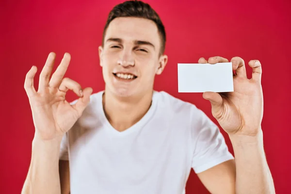 Un homme dans un T-shirt blanc avec une carte de visite dans ses mains émotions fond rouge — Photo