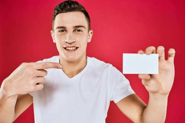 Un homme dans un T-shirt blanc avec une carte de visite dans ses mains émotions fond rouge — Photo