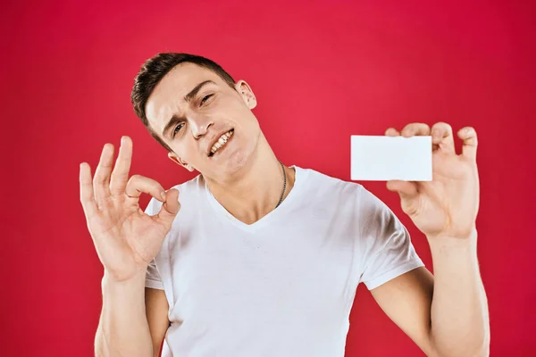 Een man in een wit T-shirt met een visitekaartje in zijn handen emoties rode achtergrond — Stockfoto