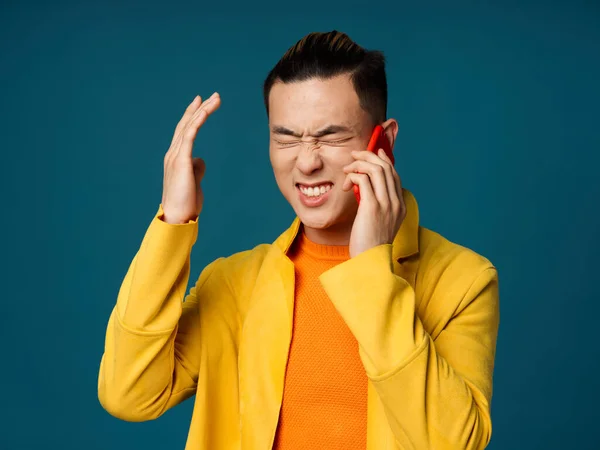 Hombre asiático haciendo un gesto con las manos y hablando por teléfono en un suéter azul chaqueta amarilla de fondo — Foto de Stock