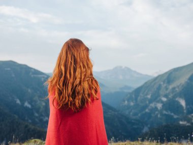 Bir kadın doğadaki dağlara ve omuzlarında kırmızı bir ekoseye bakar.