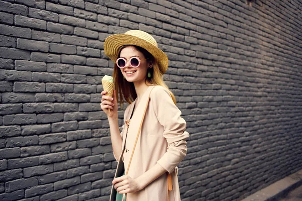 Женщина в шляпе в солнцезащитных очках, мороженое проходит через городскую кирпичную стену — стоковое фото