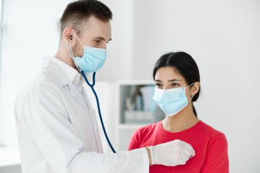 Steteskoplu bir doktor, tıbbi maske takan bir hastayı muayene ediyor.