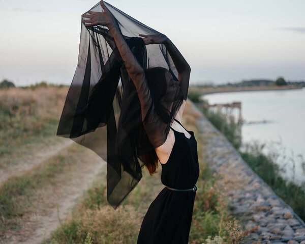 Romantic woman in nature in a black dress with a transparent scarf over her head. High quality photo