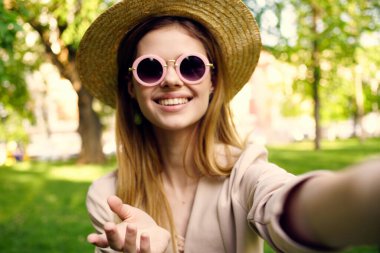 Güneş gözlüklü ve şapkalı neşeli kadın parkta yeşil çimenler üzerinde yürüyor.