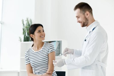 Beyaz önlüklü erkek doktor, bir kadının eline sağlık hastanesi aşısı enjekte ediyor.