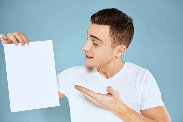 Een man in een wit t-shirt houdt in zijn handen een vel papier emoties bijgesneden weergave van een blauwe achtergrond Copy Space — Stockfoto