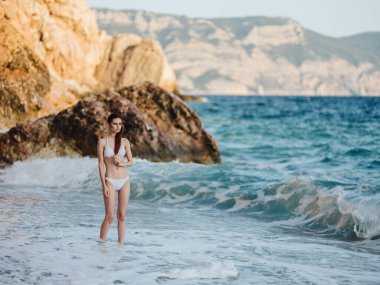 Deniz kenarında mayo giymiş romantik bir kadın. Beyaz köpüklü plaj ve arka planda kayalar.