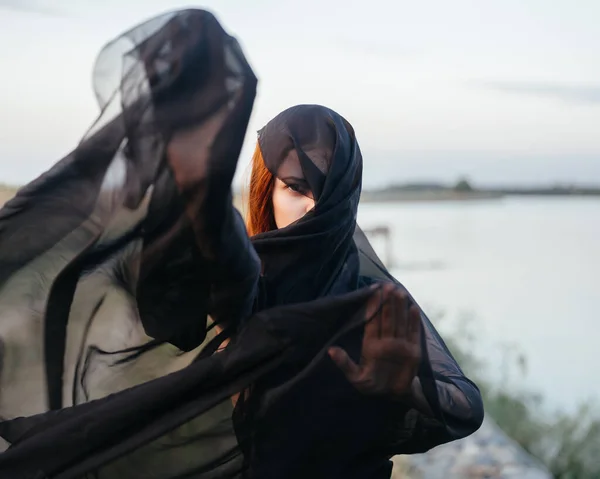 Uma mulher com um lenço preto no rosto perto do rio na natureza — Fotografia de Stock