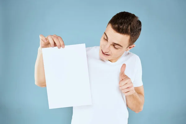 Een man in een wit t-shirt houdt in zijn handen een vel papier emoties bijgesneden weergave van een blauwe achtergrond Copy Space — Stockfoto