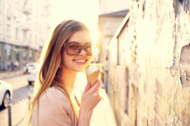 Şehirde güneş gözlüklü neşeli bir kadın dondurma ile sokakta yürüyor.