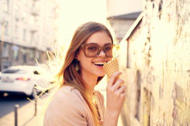 Güneş gözlüklü neşeli kadın elinde dondurmayla dışarıda yürüyor.