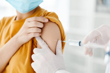 Tıbbi maske takan bir hastanın omzuna aşı enjekte eden doktorun yan görüntüsü. 