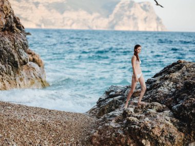 Beyaz mayo giymiş güzel bir kadın doğada yüksek bir taşın üzerinde duruyor ve beyaz köpüklü okyanus berrak sularda.