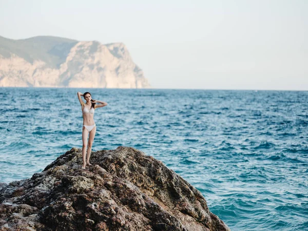 Beautiful woman in a white swimsuit stands on a high stone in nature and white foam ocean clear water — Stock Photo, Image