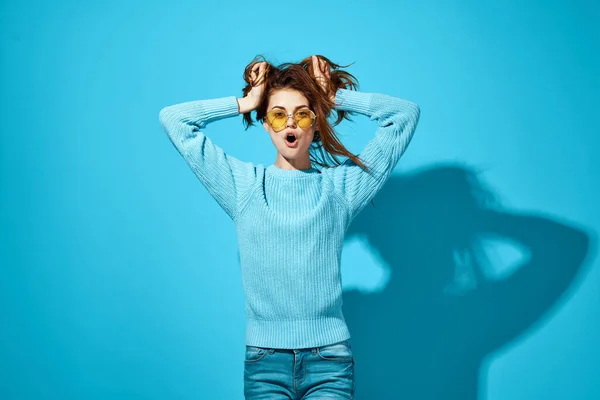 Bela mulher em azul suéter amarelo óculos de sol sorriso segurando cabelo estúdio azul fundo — Fotografia de Stock