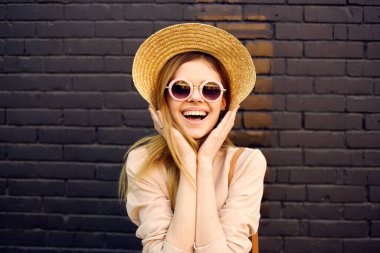 Güneş gözlüklü ve dekorasyon şapkalı bir kadın arka planda tuğla duvarda geziniyor.