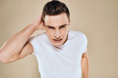 Yüzünde hoşnutsuzluk ifadesi olan beyaz tişört jestleri ve bej arka planı olan bir adam.