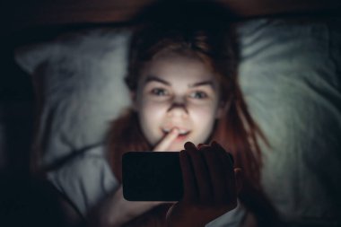 Ellerinde telefon olan duygusal bir kadın gece bağımlılığında yatakta yatıyor.