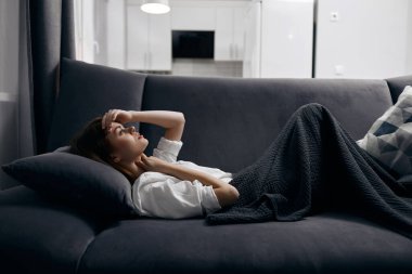 Rahat bir odada, gri bir battaniyeyle kaplı bir kanepede yatan bir kadın.