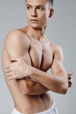 Kol kasları şişmiş atletik bir adam kollarıyla kendine sarılıyor ve gri bir arka plana bakıyor.