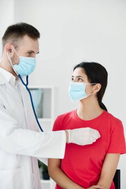 Steteskoplu bir doktor tıbbi maske takan bir hastayı muayene ediyor.