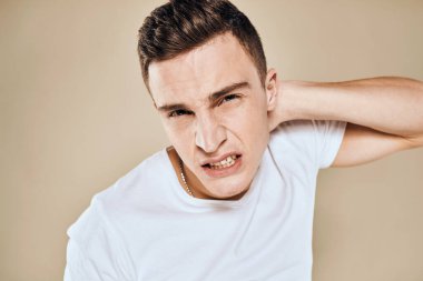 Yüzünde hoşnutsuzluk ifadesi olan beyaz tişört jestleri ve bej arka planı olan bir adam.