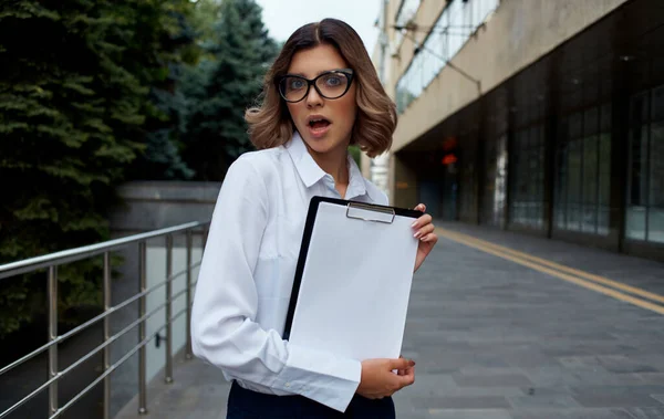 Happy business woman with documents near the building on the street in glasses and in a white shirt