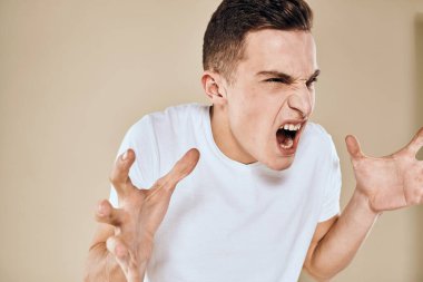 Yüzünde hoşnutsuzluk ifadesi olan beyaz tişört jestleri ve bej arka planı olan bir adam.