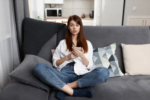 Mulher surpreso com um telefone em suas mãos sentado em um sofá confortável em casa estilo de vida — Fotografia de Stock