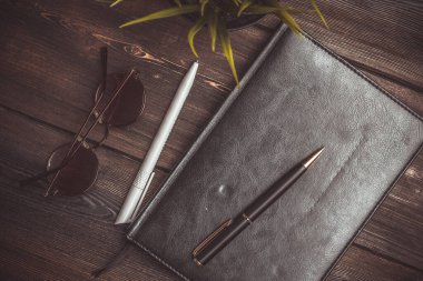 Siyah kaplı bir kitap ve tahta bir masanın üzerine uzanmış kalem işleri çiçek saksısı kahve fincanı.