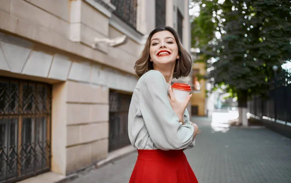 Mujer con una taza de café en la mano cerca de un edificio de ladrillo y verano maquillaje falda roja — Foto de Stock