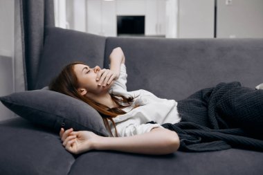 Kadın, ev sağlığı sorunlarında kanepede yatarken elleriyle yüzünü kapatıyor.