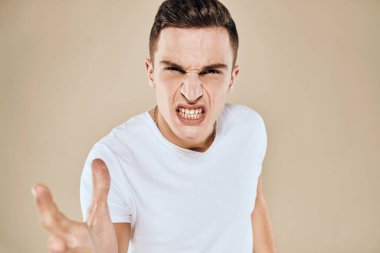 Yüzünde hoşnutsuzluk ifadesi olan beyaz tişört jestleri ve bej arka planı olan bir adam.