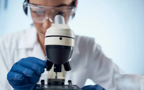 Asistente de laboratorio femenino examina las bacterias a través de un microscopio y gafas en la vacunación del elemento químico de la cara — Foto de Stock