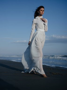 Okyanus kıyısında beyaz elbiseli romantik bir kadın kumların üzerinde yalınayak yürüyor.