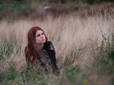 Siyah elbiseli kızıl saçlı kadın, kuru çimlerin üzerinde bir tarlada yatıyor. 