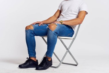 Kotlu ve tişörtlü bir adam arka planda bir sandalyede oturuyor.