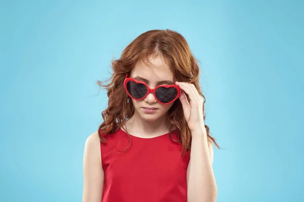 Little girl in sunglasses in the form of hearts red dress curly hair blue background — Stock Photo, Image