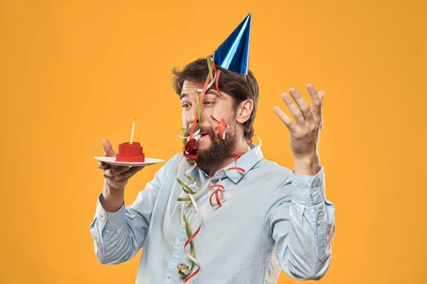 Hombre alegre con un pastel en un fondo amarillo cumpleaños días festivos gorra en su cabeza — Foto de Stock